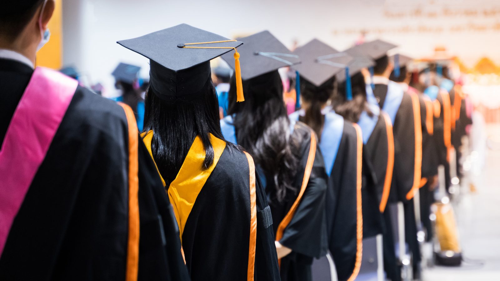 A line of graduates in caps and gowns walk down the aisle at graduation. Receiving their graduate degrees